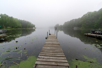 Obraz premium Looking down a pier on a calm Wisconsin lake on a foggy morning. Fishing boat in distance.