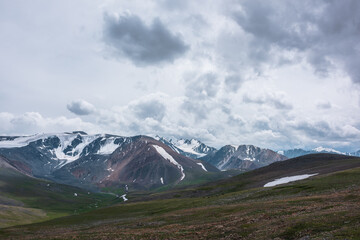 Fototapeta premium Dramatic alpine view from stony grassy hill to large snow-capped mountain range under cloudy gray sky. Beautiful mountain landscape with green rock hills against high snowy mountains in rainy weather.