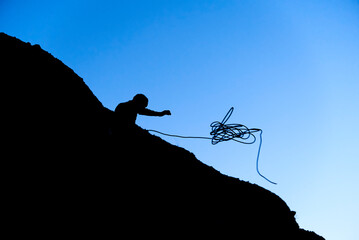 Backlit silhouette of a climber throwing a rope on a mountain ridge at sunset. Image with copy...