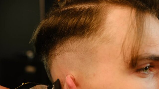 close up men's hairstyling and haircutting with hair clipper in a barber shop or hair salon. Hairdresser service in a modern barbershop in a dark key lightning with warm light side view