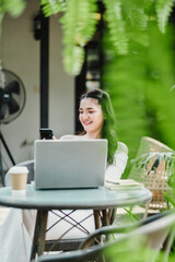 Smiling young woman enjoys checking her smartphone while seated with a laptop at a garden cafe, creating a harmonious work environment.