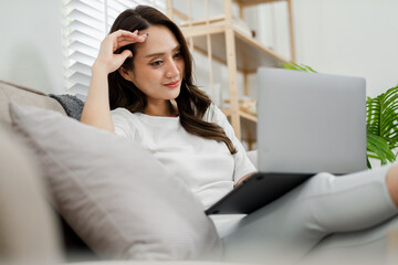 Woman reclines on a couch at home, working on a tablet with a thoughtful expression, surrounded by a peaceful indoor environment.