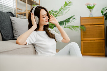 Woman closes her eyes, savoring the music through her headphones, while relaxing on a sofa in a bright, plant-filled room.