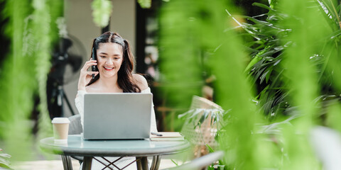 Woman is seen happily conversing on her phone while working on a laptop at a garden table, with a coffee and greenery around her.