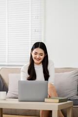 Young woman sits at a coffee table, smiling as she works on her laptop in the comfort of her sunlit living room, creating a relaxed work atmosphere.