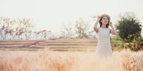 Woman in a white dress and straw hat standing amidst a golden wheat field at sunset.