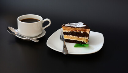 A plate with a piece of chocolate sponge cake with mint leaves and a cup of hot black coffee on a black background.