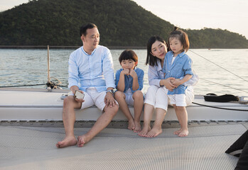 Happy Asian family relaxing, sitting, and playing on the polyester net of a luxury yacht during a sailing trip on the sea, enjoying the summer holiday concept.