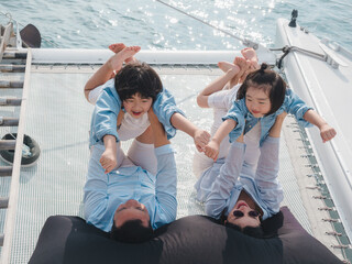 Happy Asian family relaxing, sitting, and playing on the polyester net of a luxury yacht during a sailing trip on the sea, enjoying the summer holiday concept.