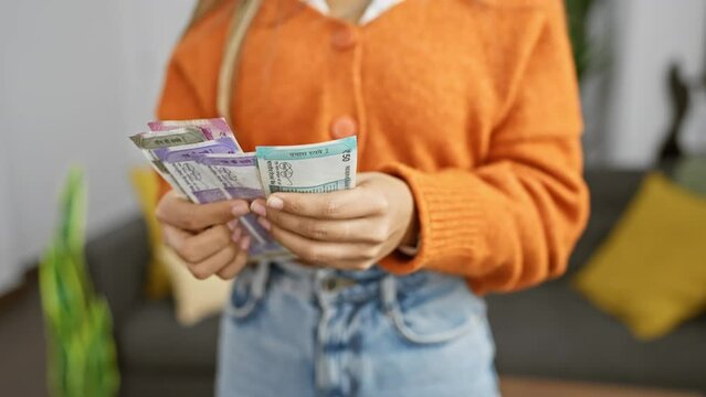 A young woman in an orange sweater counts indian rupees in her cozy indoor living space.