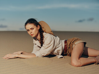 Woman laying on sandy desert with hands on hips and hat on head under clear blue sky