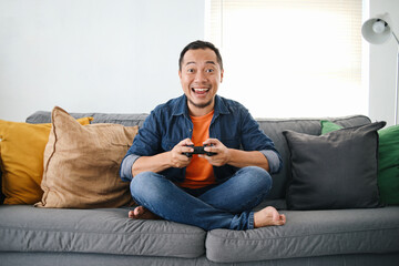 Happy Asian man playing video games with wireless joystick while sitting on comfortable sofa at home