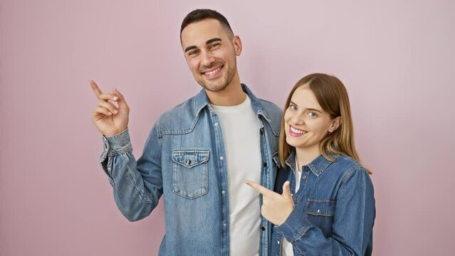 Cheerful beautiful couple in denim shirts pointing up, with natural smiles over pink isolated background. happy expression captured, man and woman standing side by side in love.