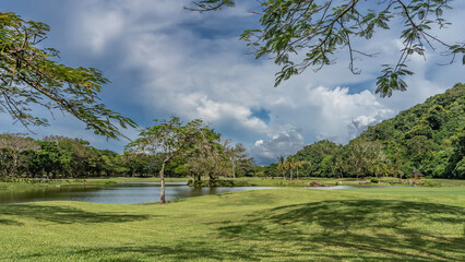 A golf course in a park. Tropical trees and palms grow on the neatly trimmed lawn. A calm lake and a gazebo for relaxing in the distance. A hill against a blue sky and clouds. Malaysia. Borneo. 