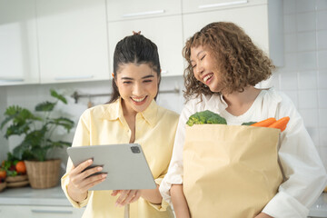 Two smiling women in a modern kitchen, one holding a grocery bag with vegetables while the other uses a tablet.