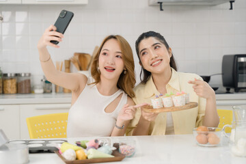 Two Asian women in a modern kitchen taking a selfie with a plate of beautifully decorated cakes, both smiling happily.