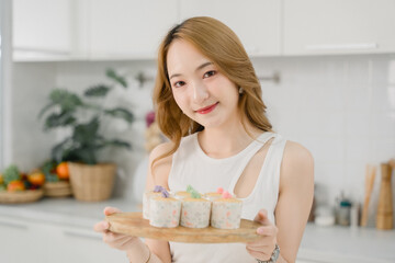 Cheerful Asian woman presenting a tray of colorful handcrafted cupcakes in a bright, contemporary kitchen.