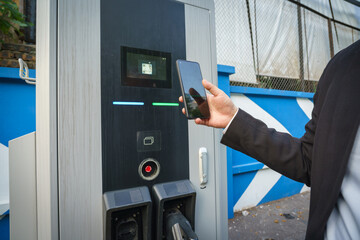 Man holding smartphone with application to charging car at electric vehicle charging station in...