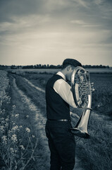 A young man in a white shirt stands on a dirt road with a trumpet - tenor saxhorn