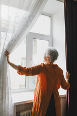 Back view of stylish gray-haired senior lady in orange home dress opening drapes to let light in, greeting new good day, staring at vigorous city life outside, looking into future with hope