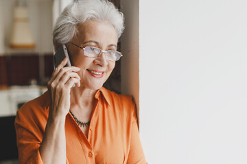 Indoor image of smiling aged female with gray hair in glasses talking on phone standing near window, having nice conversation with her bestie, planning to go for shopping on weekend together