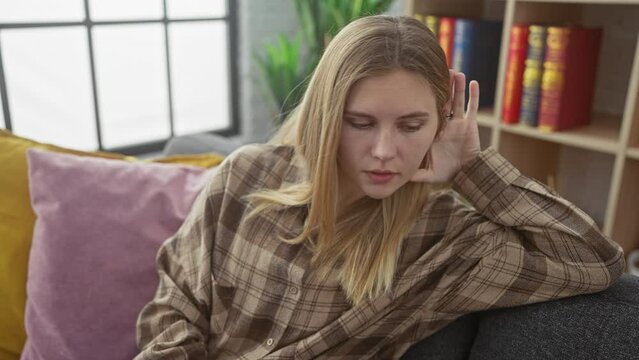 At home, a happy young blonde woman, hand over ear, sitting cozy on her sofa, drinking in juicy gossip and rumors, despite deafness problems.