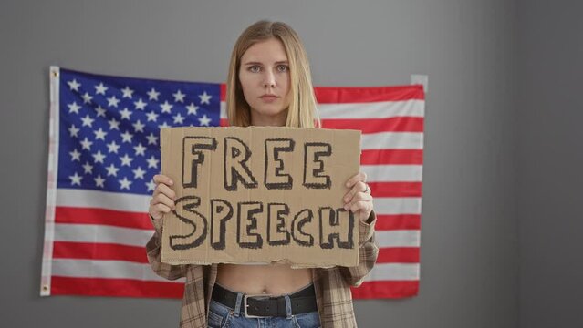 A serious blonde woman holding a 'free speech' sign in front of an american flag indoors