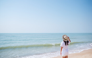 Rear view image of a young woman with hat standing on the beach with blue sky background