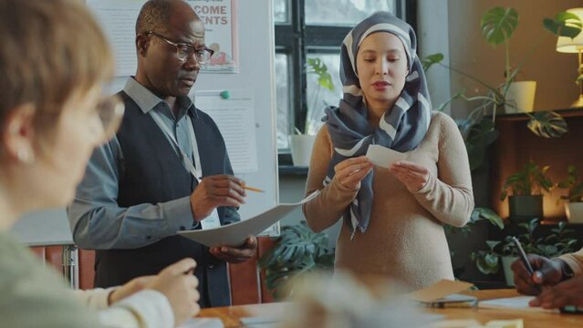 Young female student in hijab showing educational cards and explaining new English words in front of African American teacher and group mates during language class in immigrant school