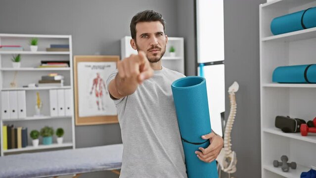 Confident young hispanic man pointing at you from a rehab clinic, holding a yoga mat, making a choice, all serious and full of energy
