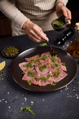 tuna carpaccio - woman puts capers green sauce onto slices of fresh raw tuna fillet on black ceramic plate