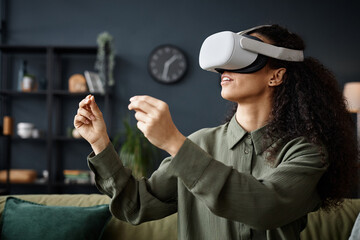 Medium closeup of young woman wearing VR headset sitting on sofa in living room playing video game...