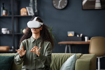 Young woman wearing VR headset sitting on sofa in living room typing text in the air, copy space