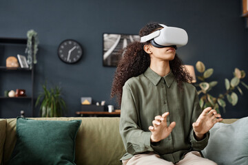 Medium shot of young biracial woman wearing VR headset sitting on couch in living room working...