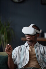 Vertical shot of young African American man wearing VR headset sitting in living room writing text...