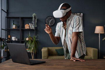 Medium long shot of young African American man wearing VR headset standing in living room looking...