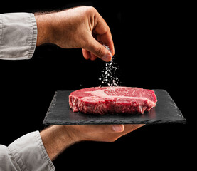 Chef is salting or seasoning raw ribeye steak laying on graphite serving board. Black background.