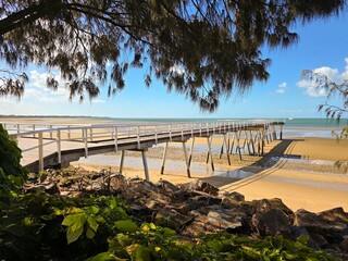 Calm beach at low tide with clear blue sky, yellow sand, a long pier and gumtrees.