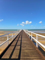 A long wooden pier stretching to the horizon, over a calm beach at low tide with clear blue sky, yellow sand and little white clouds.