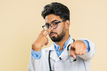 I am watching you. Confident attentive Indian young doctor cardiologist man pointing at eyes and camera spying on someone gesture intimacy. Arabian apothecary pharmacy guy isolated on beige background