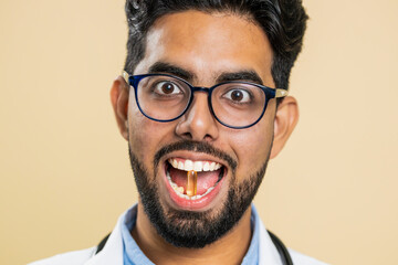 Smiling Indian doctor man puts of tablet pill vitamin D or omega-3 yellow capsule into mouth,...