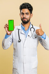 Indian doctor cardiologist man hold smartphone with green screen chroma key mock up recommend good application promotional sale offer phone number. Arabian apothecary guy on beige background. Vertical