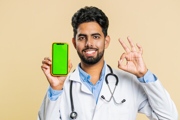 Indian young doctor cardiologist man hold smartphone with green screen chroma key mock up recommend...