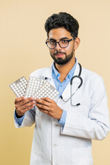 Indian doctor cardiologist man shows lot of tablet pills in blisters, recommends drugs vitamin antibiotic cure treatment. Health care. Arabian apothecary pharmacist guy on beige background. Vertical