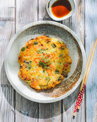 A Korean vegetable pancake garnished with fresh herbs on a speckled ceramic plate against a textured gray background.