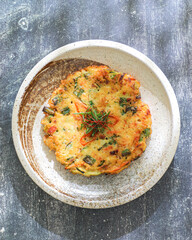 A Korean vegetable pancake garnished with fresh herbs on a speckled ceramic plate against a textured gray background.