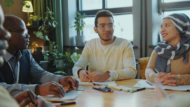 Group of diverse immigrant students and senior African American teacher sitting at table in classroom and having conversation as practicing English speaking skills in language school