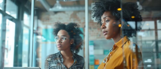 Obraz premium In an agency meeting room, two diverse multiethnic women discuss business results on laptops and app designs behind glass walls.