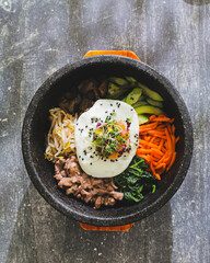 A vibrant Korean bibimbap served in a black stone bowl, featuring sections of minced meat, various julienned vegetables, a fried egg on top, sprinkled with sesame seeds