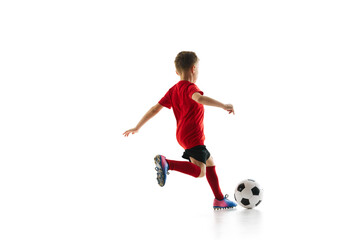 Sporty little boy, kicks ball to make perfect shot in motion against white studio background. Small football player makes perfect pass. Concept of professional sport, championship, youth league, hobby